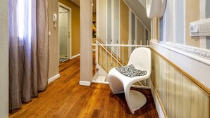 Un interior moderno con piso de madera, una silla blanca con cojín estampado, una escalera con barandas blancas y una lámpara fijada a la pared.