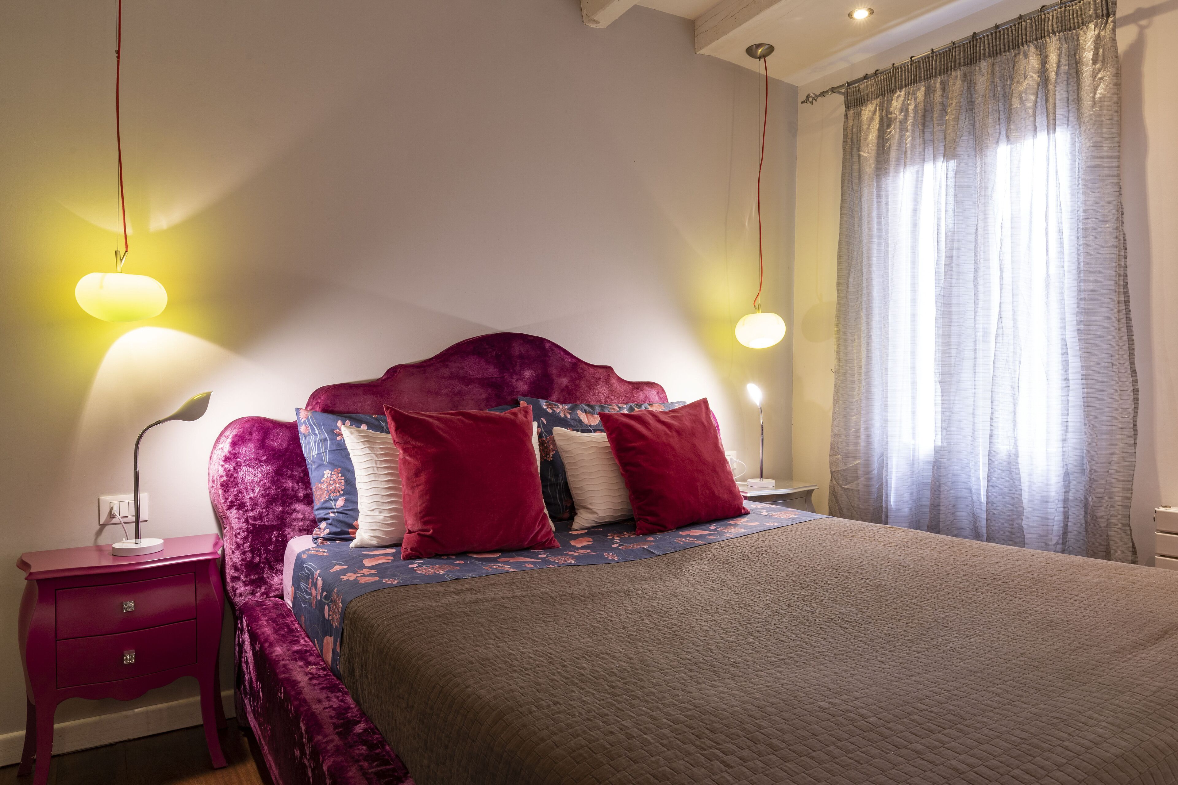 Une chambre à coucher avec un grand lit, une table de chevet, une lampe et une fenêtre avec des rideaux.