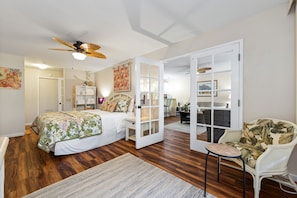 Relax in this spacious room with a large bed, comfortable chair, and dining area, featuring a ceiling fan and a door leading to another room.