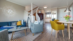 A modern living room with a blue sofa, a gray armchair, and a yellow chair. There is a dining table with a vase of flowers. - Vacation home Backbord with roof terrace, directly on the water with Baltic Sea & Schlei view (Kappeln)