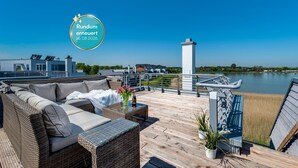 A rooftop deck with a wicker sofa, a small table with a vase of flowers, and a view of a lake. - Vacation home Backbord with roof terrace, directly on the water with Baltic Sea & Schlei view (Kappeln)