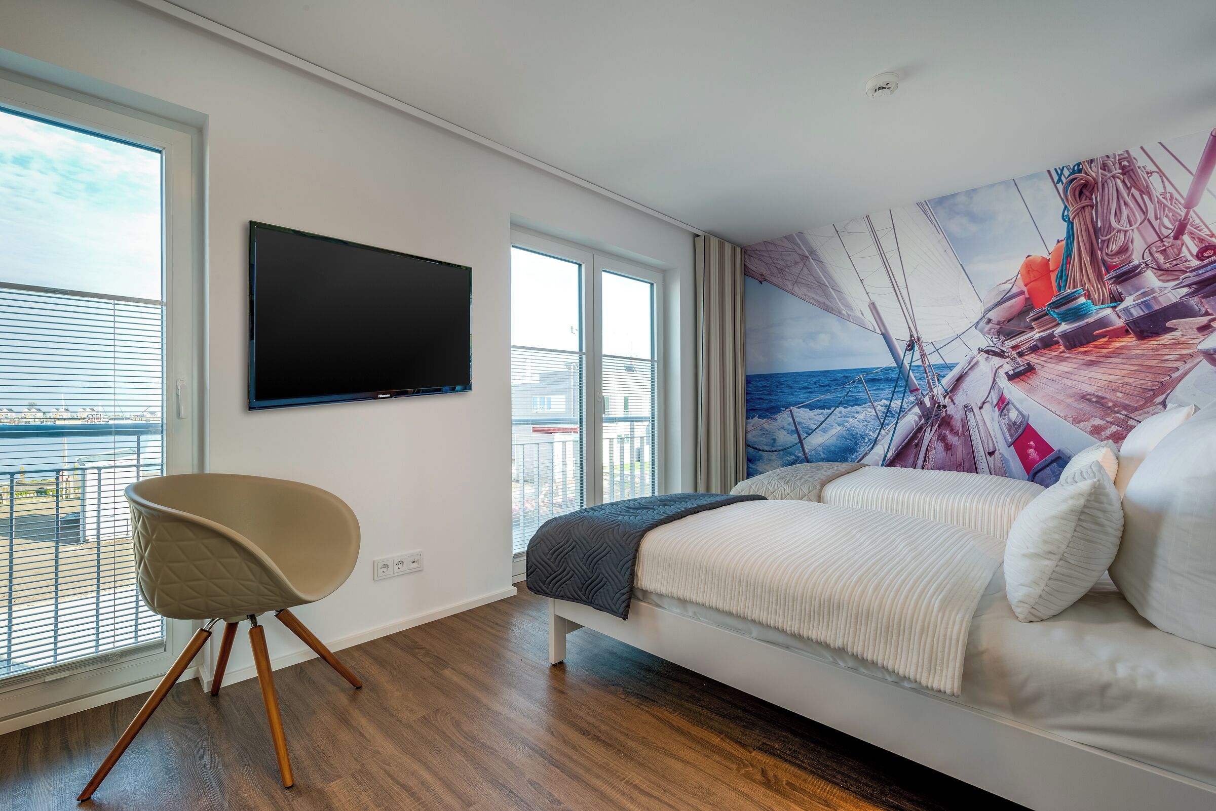 Een moderne slaapkamer met een groot raam, een flatscreen televisie, een bed met witte beddengoed en een muurschildering van een zeilboot.