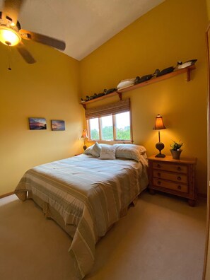 A bedroom with a bed, bedside tables, a ceiling fan, a window with curtains, and wall-mounted shelves with decorative items.