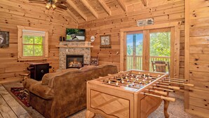 Acogedor interior de cabaña de madera con una mesa de fútbol de mesa, chimenea, televisor y un ventanal grande.
