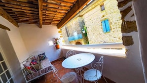 Un patio acogedor con una mesa redonda y sillas, un banco acolchado y vistas a un edificio de piedra con contraventanas azules.