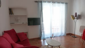 Una sala de estar con un sofá rojo, una mesita redonda pequeña, un televisor de pantalla plana y una lámpara de pie.
