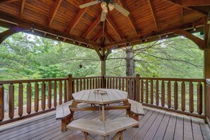 Um deck de madeira com mesa e bancos, cercado por corrimão, com vista para uma área arborizada.