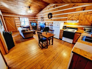 A cozy cabin interior with a wooden floor, a dining area with a table and chairs, a kitchen with a stove and refrigerator, and a living area with a sofa and TV.