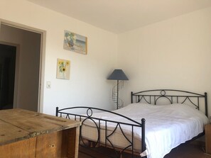 Quarto com estrutura de cama de metal, mesa de madeira, luminária e duas pinturas penduradas na parede.