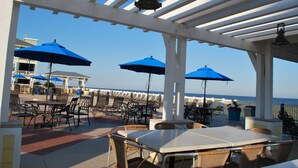 Une terrasse extérieure avec des tables et des chaises, des parasols bleus et une vue sur l’océan.
