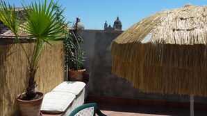 Una terraza en la azotea con un toldo de paja, plantas en macetas y vistas a edificios lejanos.