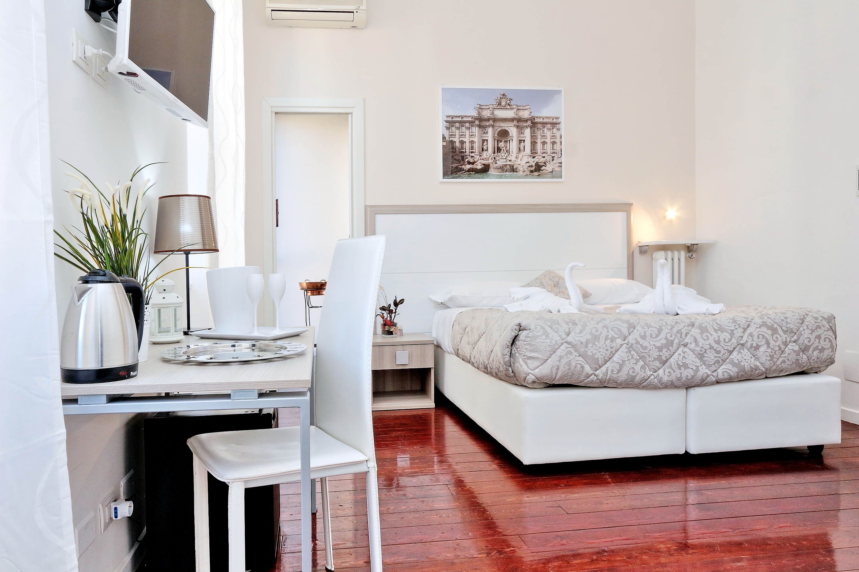 A modern hotel room with a white bed, a desk with a laptop, a chair, a potted plant, and a framed picture on the wall.