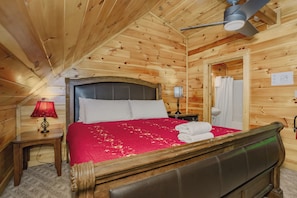 A wooden cabin bedroom with a bed, a nightstand, a lamp, a ceiling fan, and a bathroom door. - Makin Waves: Pool, S/Secluded, Pets, Games, HotTub, Wifi, near attractions! (Gatlinburg)
