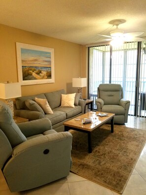 A living room with a sofa, armchairs, a coffee table, a painting on the wall, and a ceiling fan.
