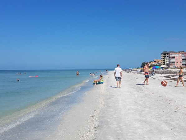A beach with clear skies, people swimming and walking, and buildings in the background.
