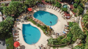 A resort with two oval-shaped swimming pools surrounded by lounge chairs and umbrellas, with palm trees and a landscaped garden.