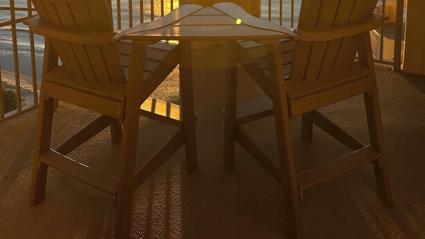 A balcony with two wooden chairs and a small table, overlooking a sunset over the ocean.