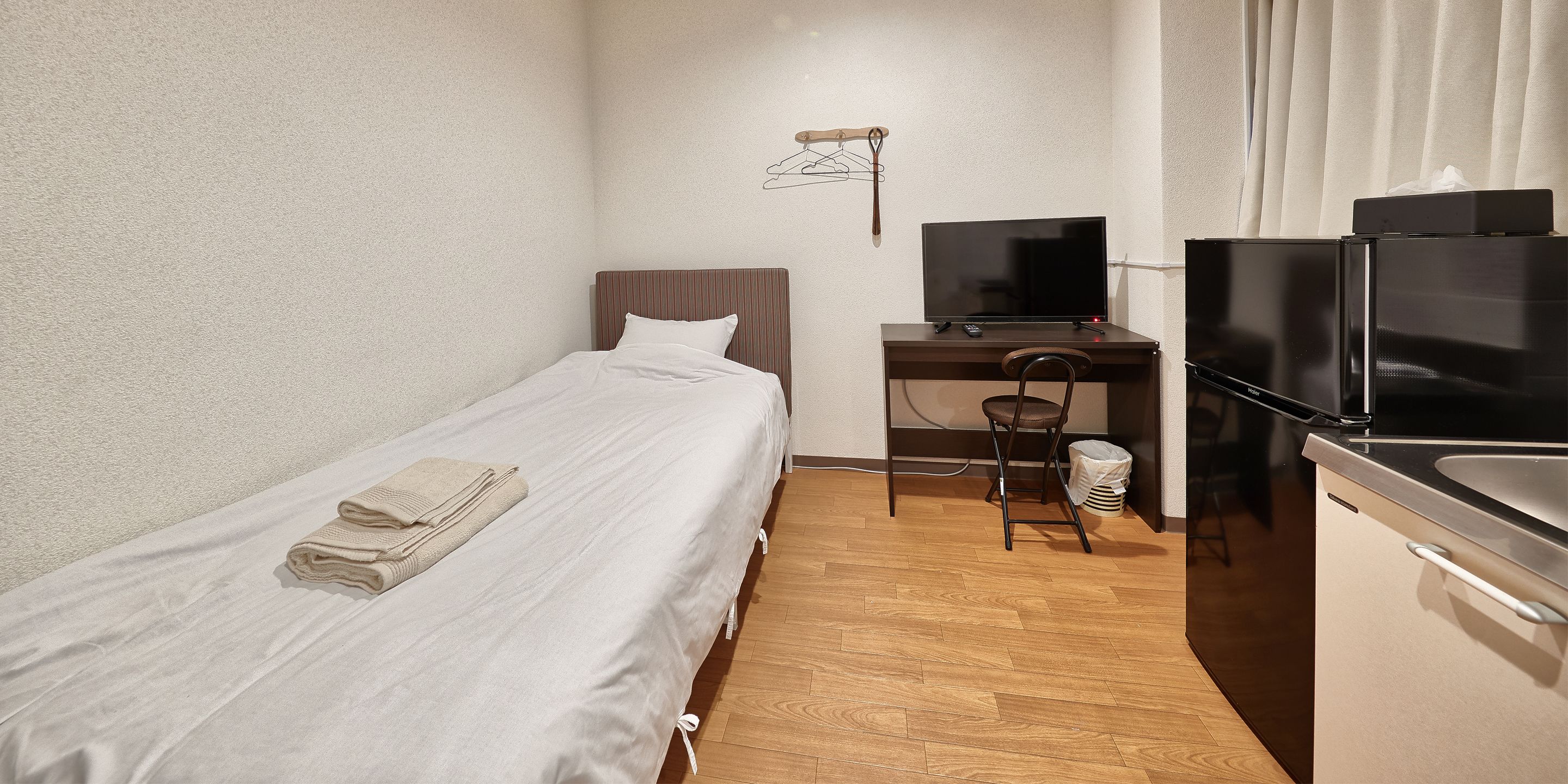 A small, neatly arranged room with a single bed, a desk with a computer and TV, and a kitchen area.