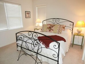 A neatly arranged bedroom with a metal bed frame, white bedding, and a red throw. There are two bedside tables with lamps, and a picture on the wall.