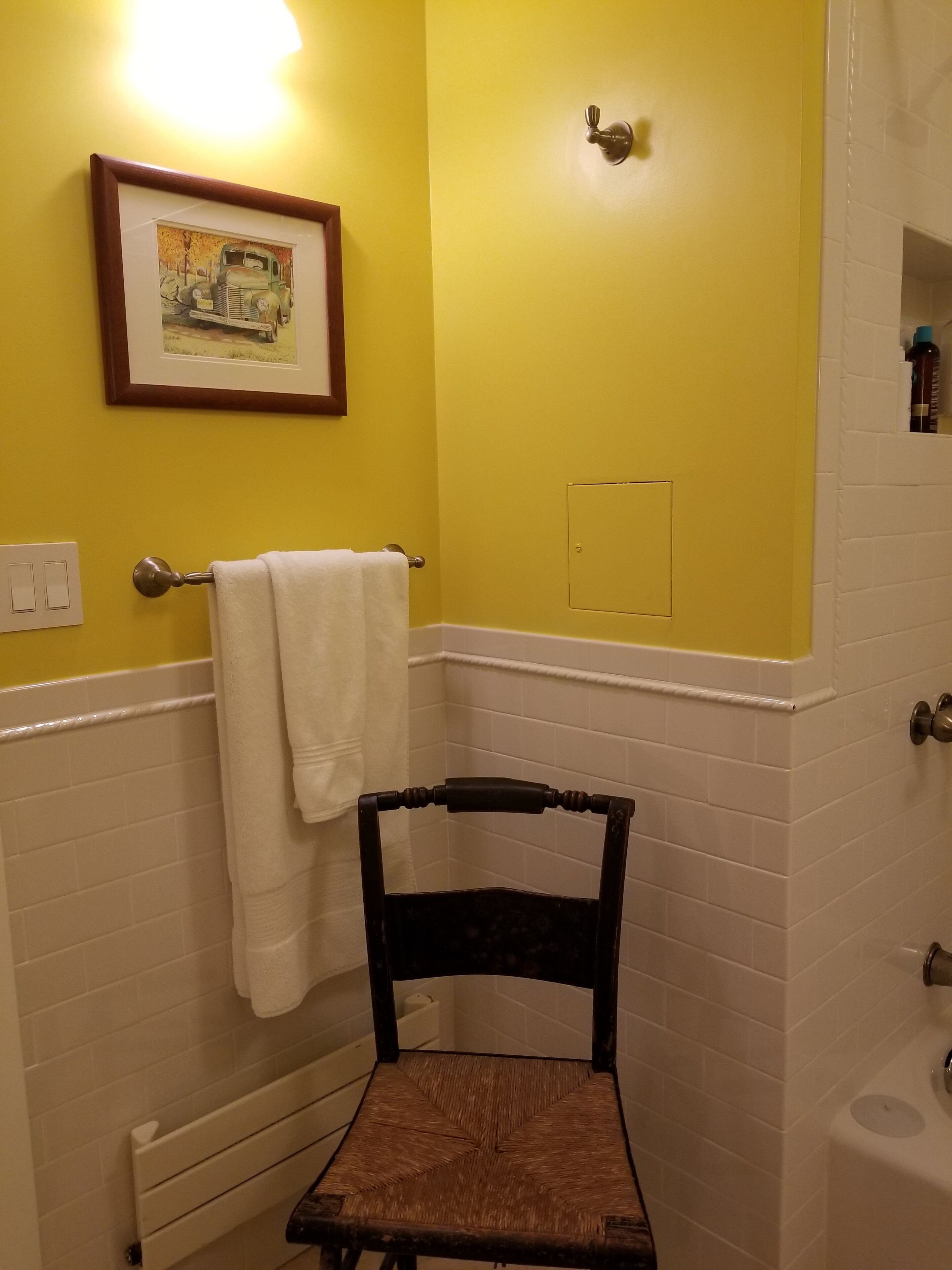 Une salle de bain avec des murs jaunes, un tableau encadré, un porte-serviettes, une chaise et une baignoire.