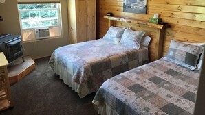 Une chambre confortable avec deux lits, une tête de lit en bois, une fenêtre donnant sur les arbres, un bureau avec un ordinateur et une petite étagère avec une boîte de mouchoirs.