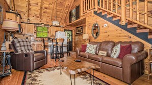 A cozy cabin interior with a wooden ceiling, a leather sofa, a dining area, and a staircase. - Enchanted Hideaway: Honeymoon, S/Secluded, Fire Pit, Hot Tub, Top Amenities, near Attractions! (Sevierville)