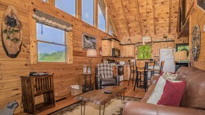 A cozy cabin interior with wooden walls, a dining area, a kitchen, and a living room with a leather couch and a coffee table. - Enchanted Hideaway: Honeymoon, S/Secluded, Fire Pit, Hot Tub, Top Amenities, near Attractions! (Sevierville)