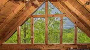 A wooden cabin with a large triangular window offering a view of a forest. - Enchanted Hideaway: Honeymoon, S/Secluded, Fire Pit, Hot Tub, Top Amenities, near Attractions! (Sevierville)
