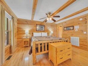 A wooden cabin bedroom with a bed, two bedside tables with lamps, a ceiling fan, a wooden bench, and a radiator.