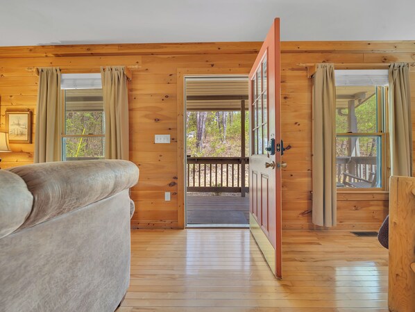 A wooden cabin interior with a bed, a door leading to an outdoor deck, and a window with curtains.