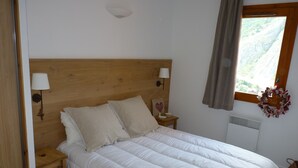 Une chambre à coucher avec une tête de lit en bois, deux tables de chevet avec des lampes, un lit avec une literie blanche, une fenêtre donnant sur un paysage montagneux et un radiateur fixé au mur.