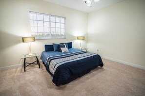 Un dormitorio con cama, dos mesitas de noche, dos lámparas, una ventana con persianas y suelo alfombrado.