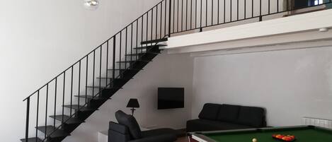 Une pièce équipée d’une table de billard, de deux canapés, d’un escalier et d’un téléviseur fixé au mur.