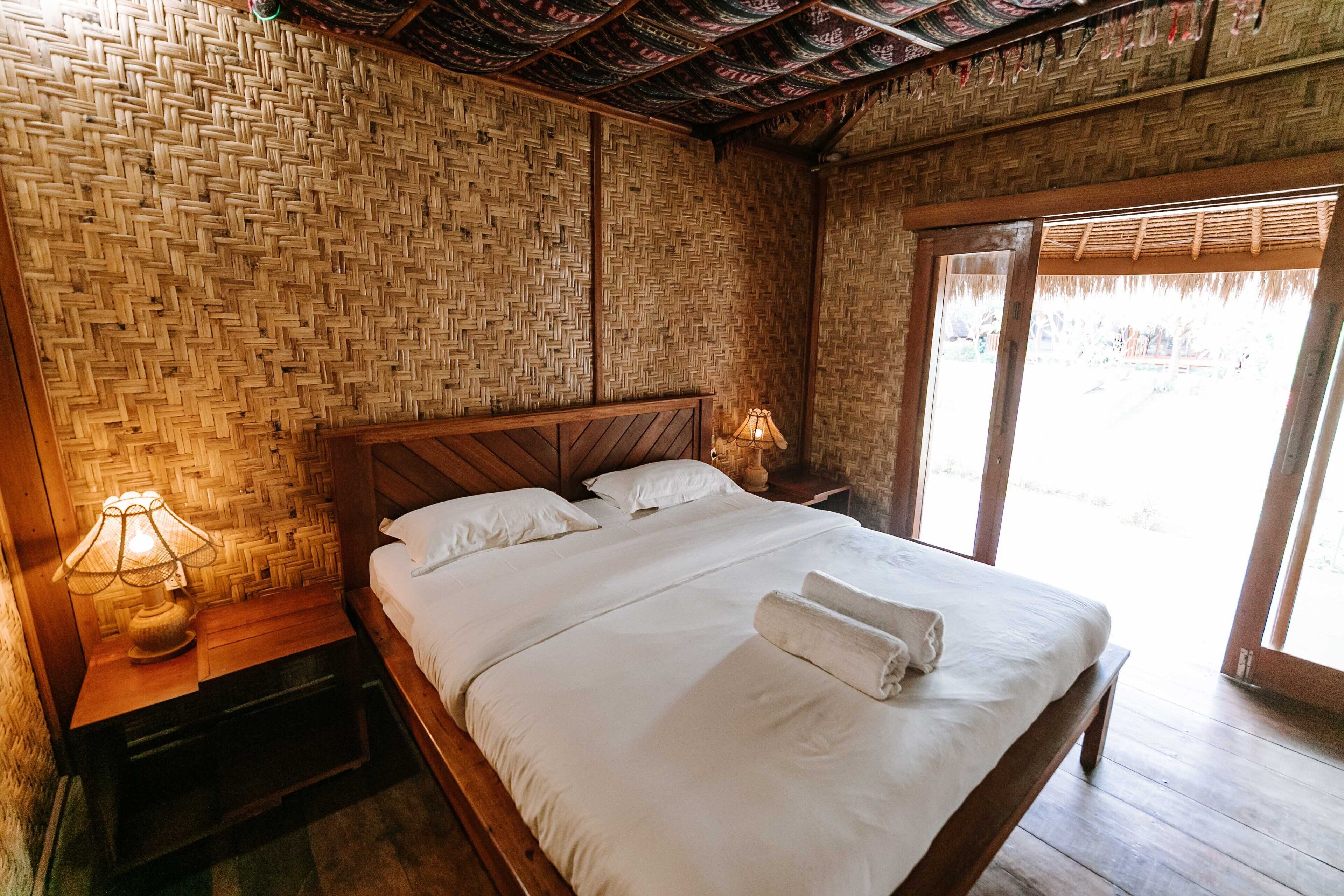 A room with a woven bamboo wall, a wooden bed with white linens, and a nightstand with a lamp.