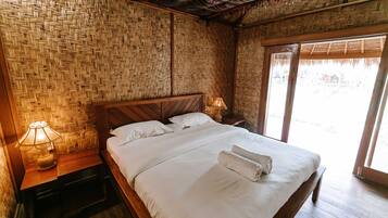 A room with a woven bamboo wall, a wooden bed with white linens, and a nightstand with a lamp.