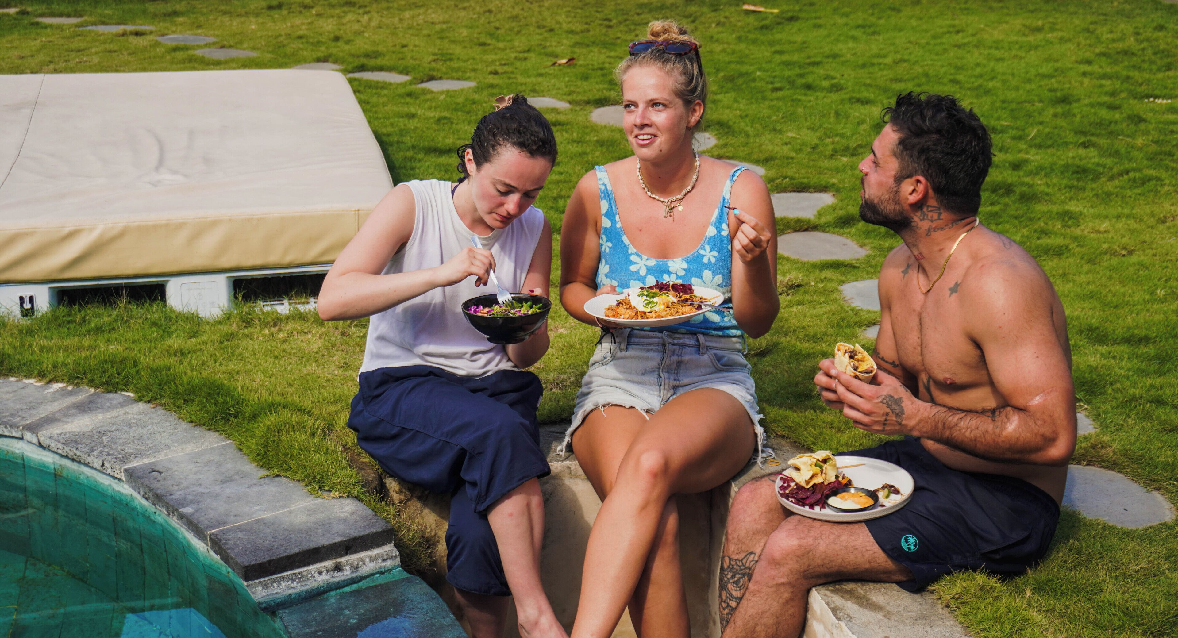 Tres personas sentadas junto a una piscina, con comida, una tienda de campaña y una zona de césped.