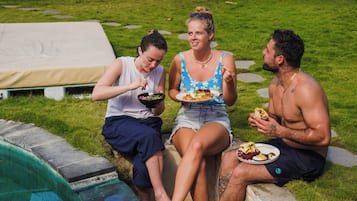 Tres personas sentadas junto a una piscina, con comida, una tienda de campaña y una zona de césped.