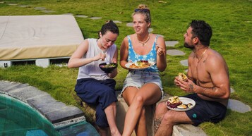 Three people sitting by a poolside with food, a tent, and a grassy area.