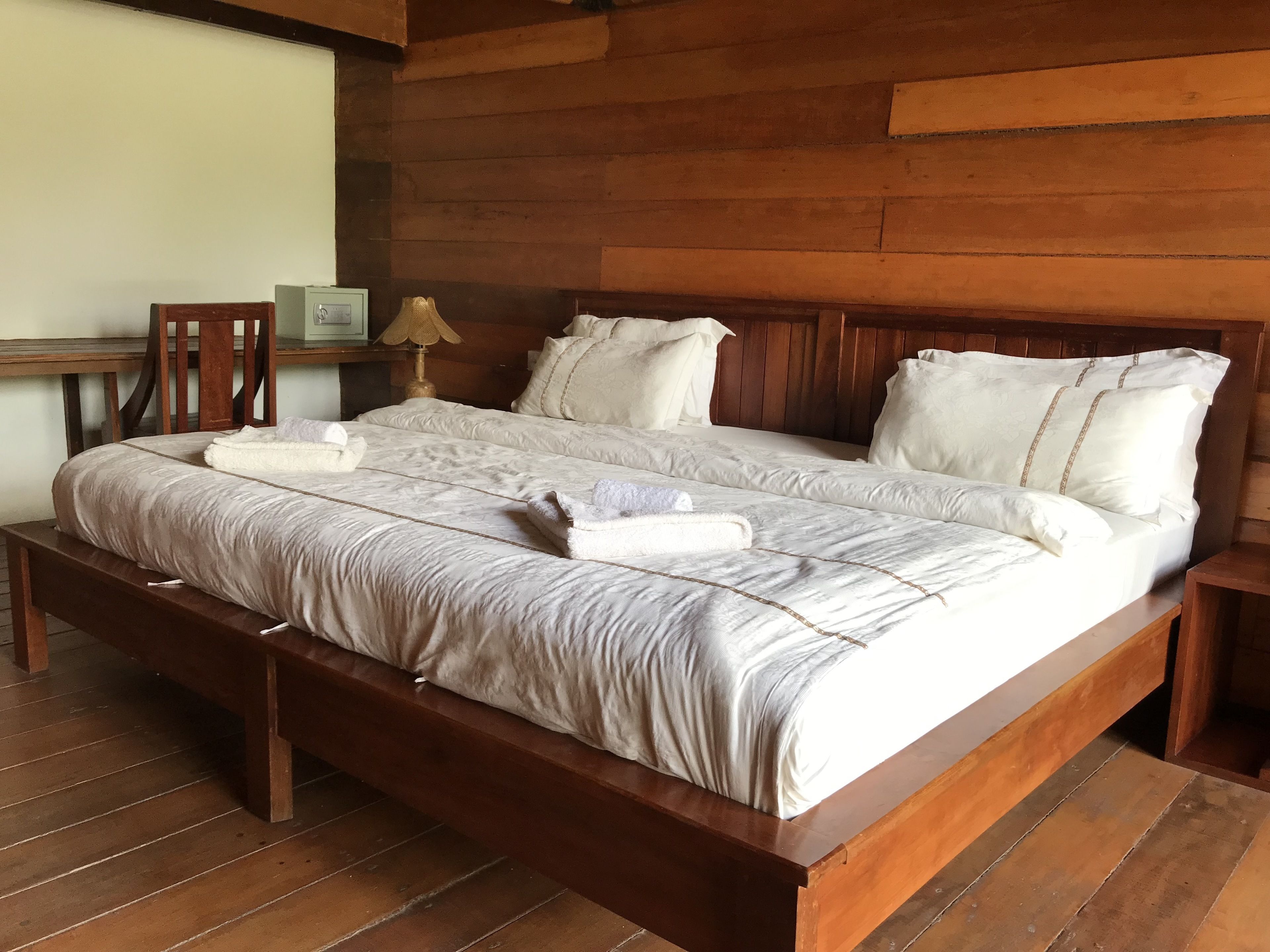 A double bed with white linens and pillows, a wooden headboard, and a wooden nightstand with a lamp.
