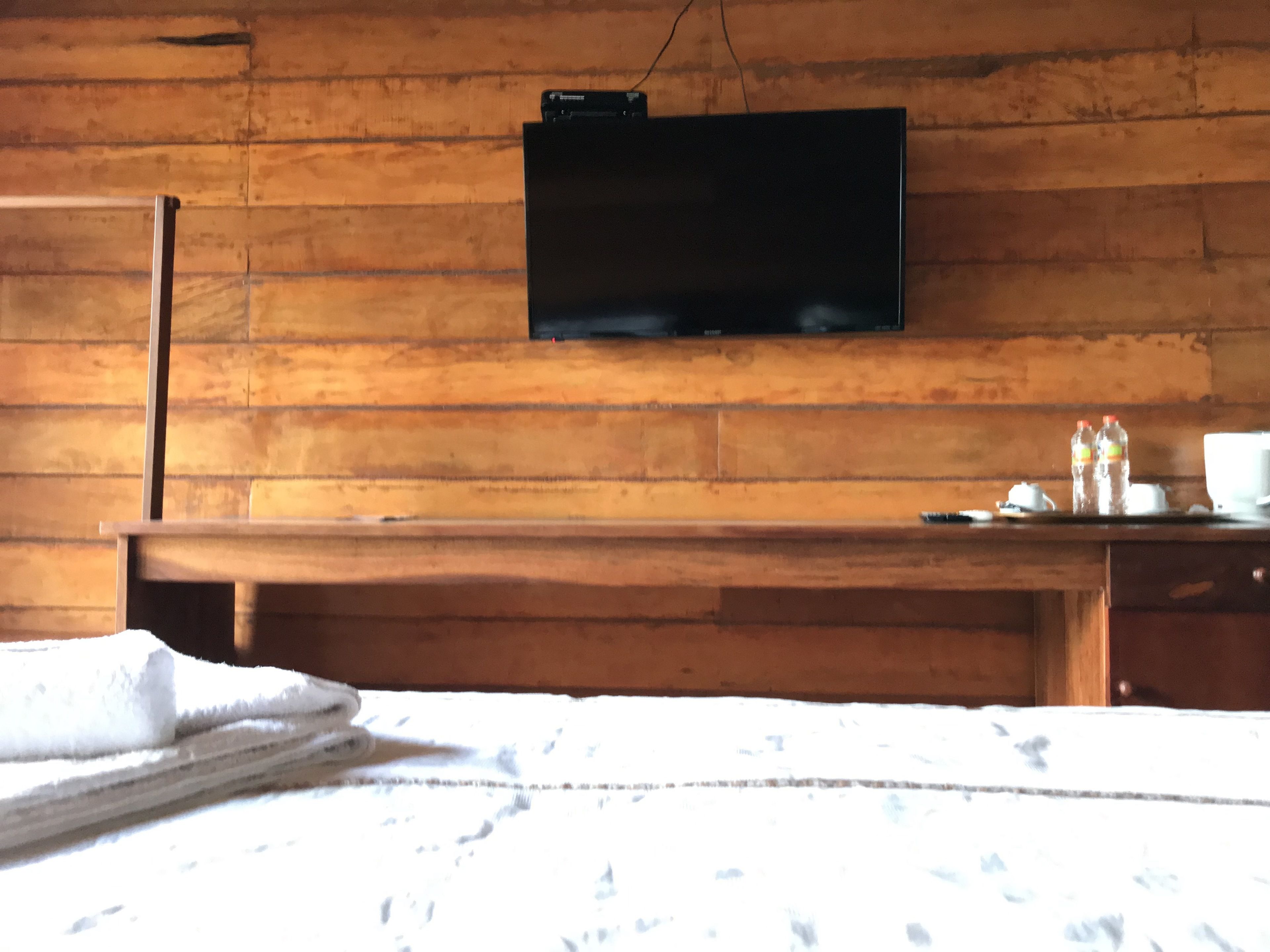 A wooden room with a bed, a flat-screen TV mounted on the wall, a wooden bench, and a small table with bottles and cups.