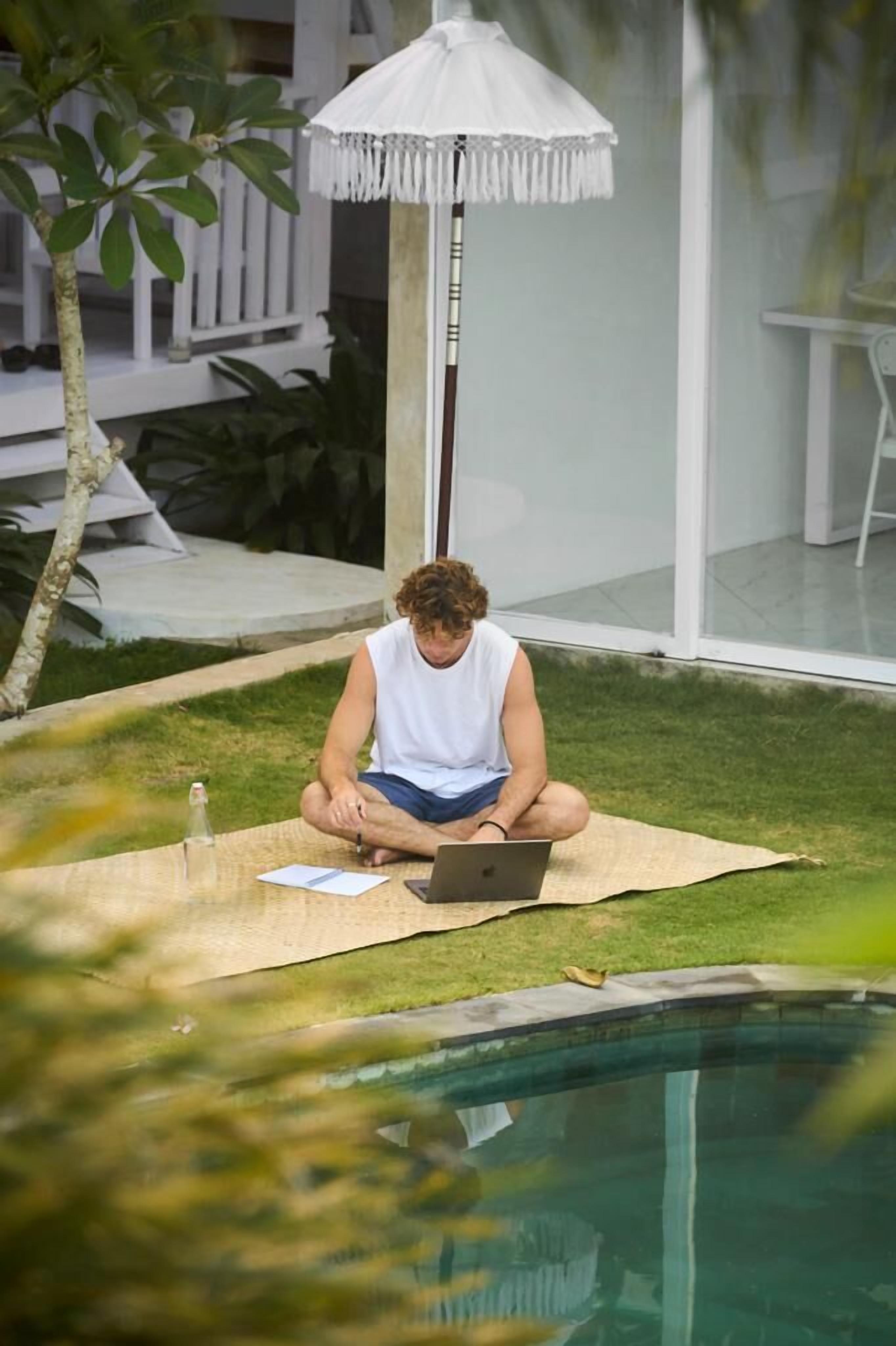 A person sitting on a mat by a pool with a laptop, under a white umbrella.