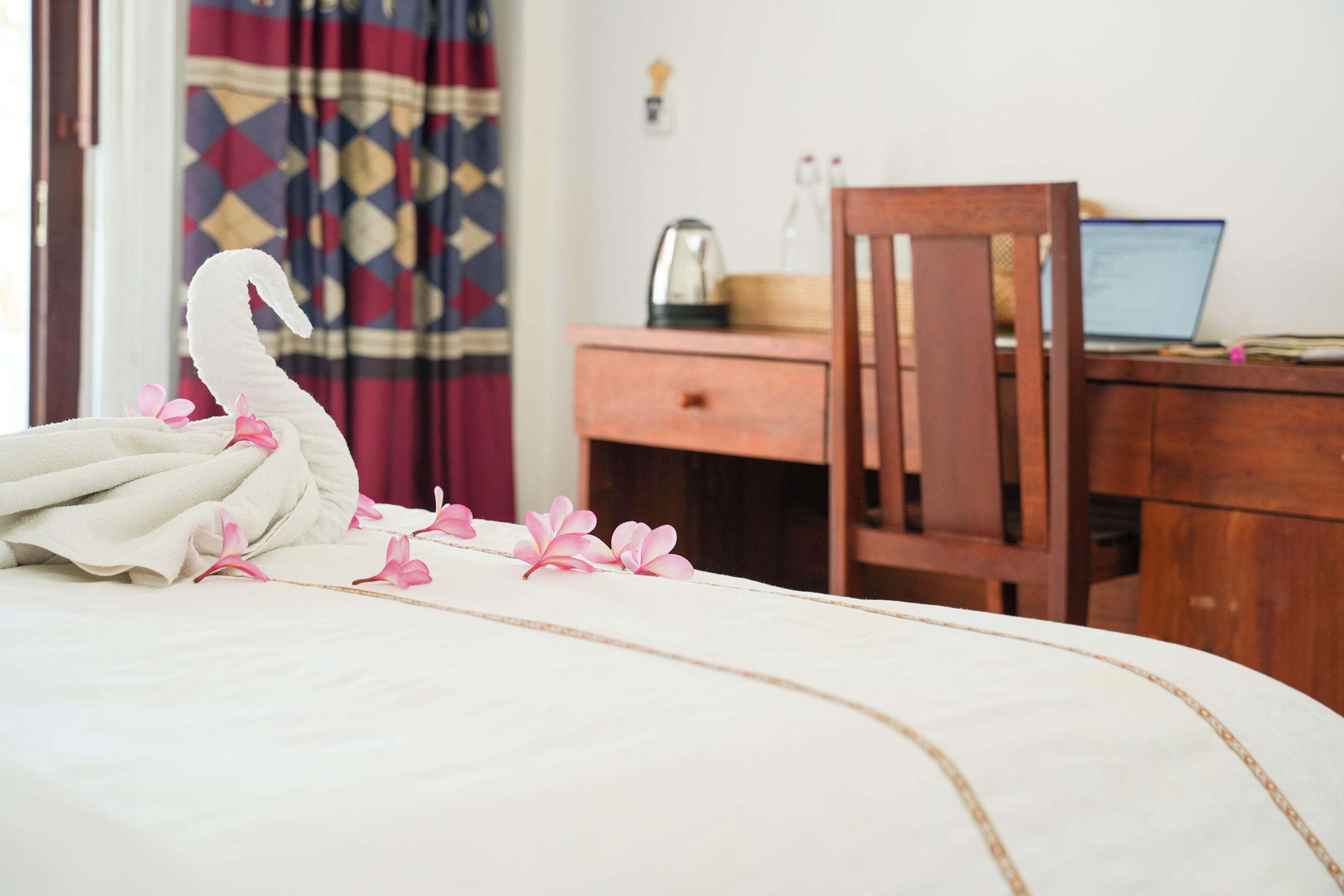 A neatly made bed with a swan-shaped pillow and pink flowers, a wooden chair, a desk with a laptop, and a patterned curtain in the background.