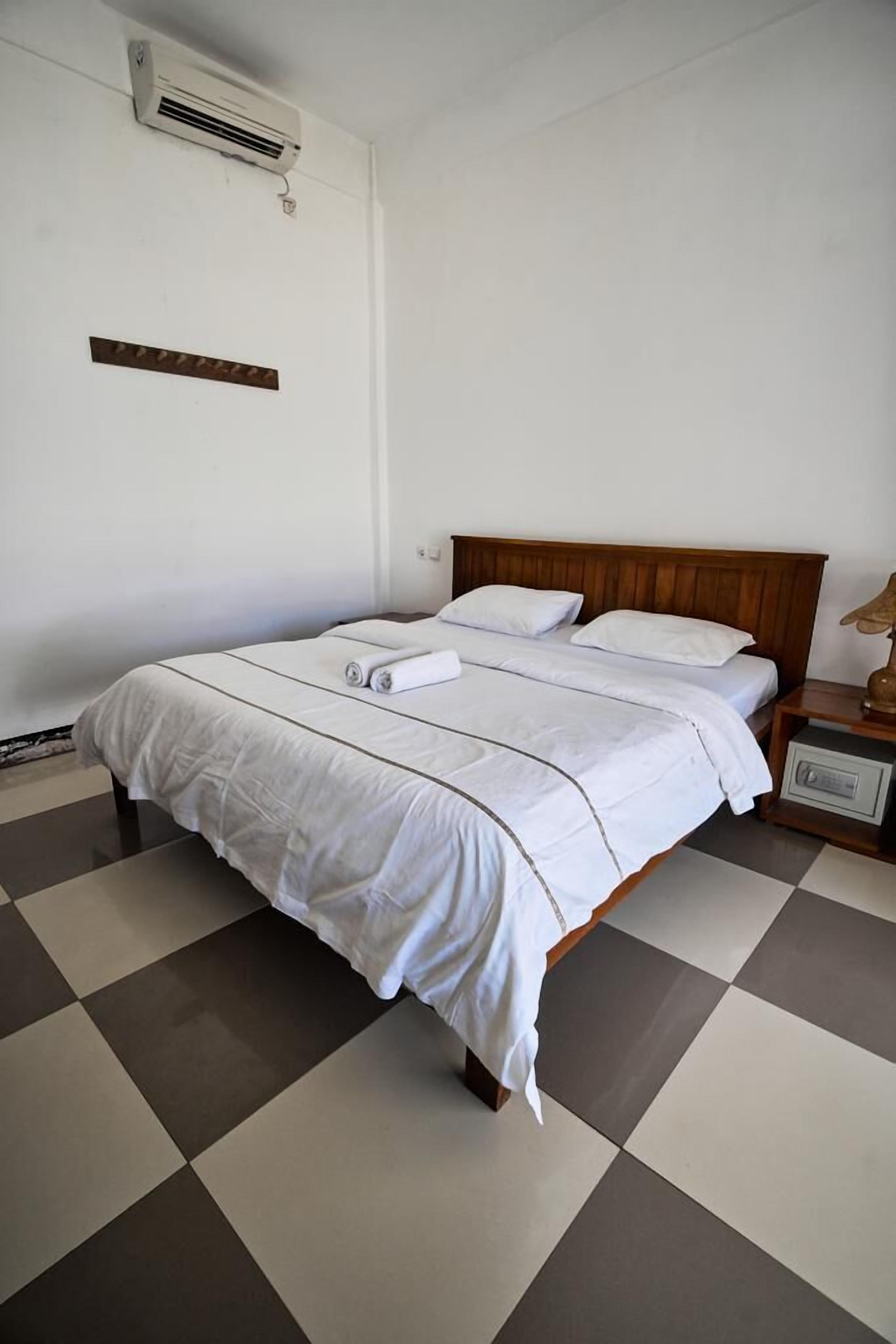 A neatly made bed with white linens and a wooden headboard, a bedside table with a lamp, and a checkered floor pattern.