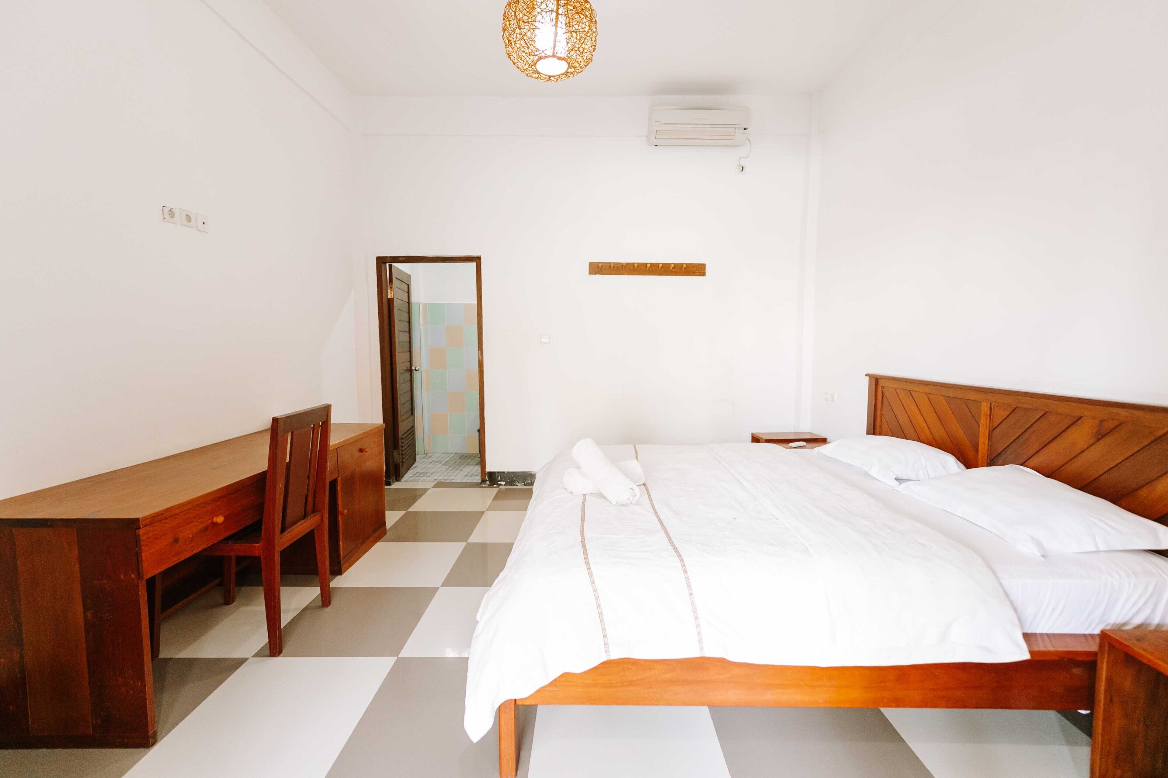 A bedroom with a wooden bed, a desk, and a chair. There is a mirror on the wall and an air conditioning unit above the bed.