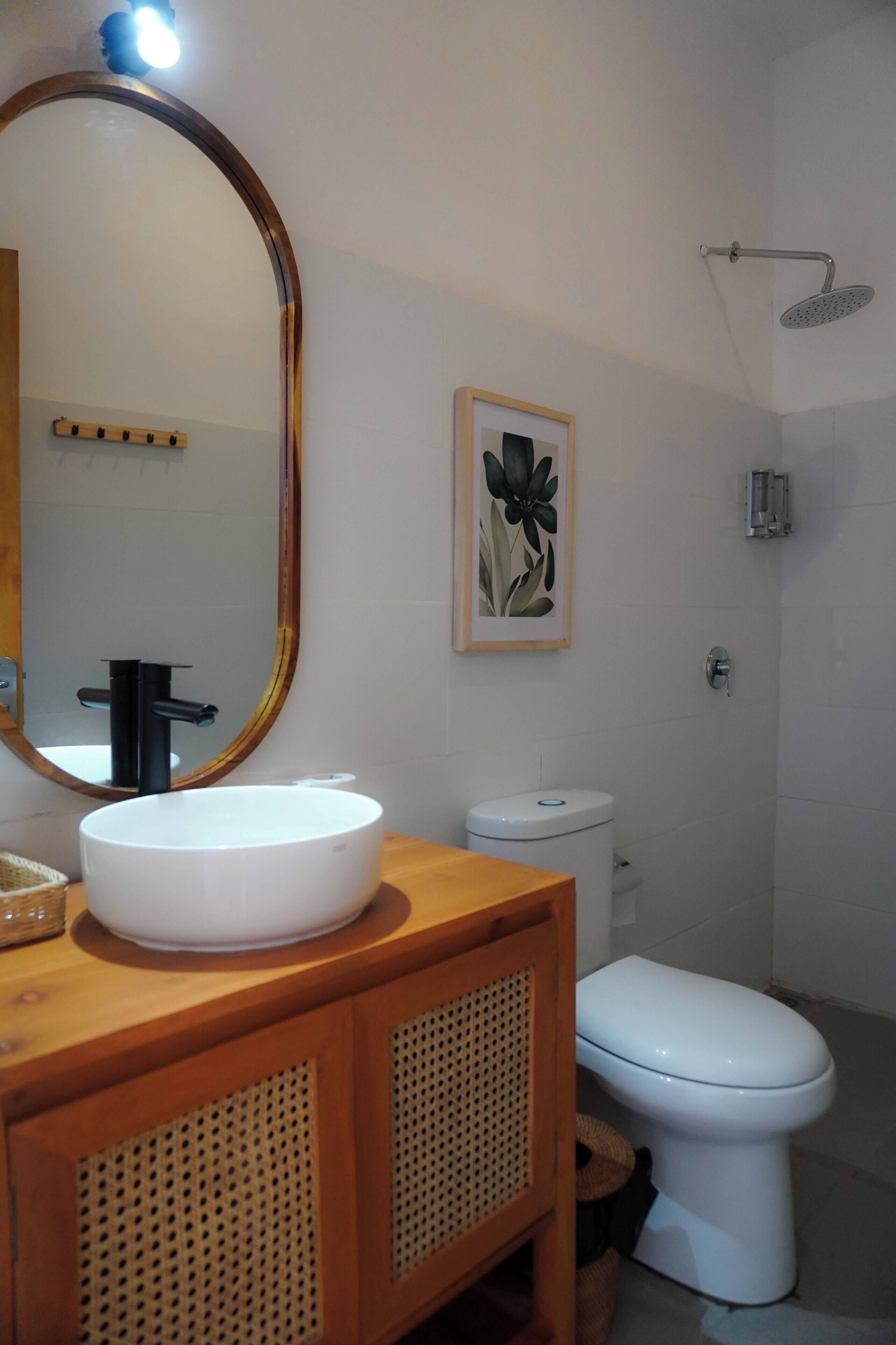 A bathroom with a wooden vanity, a white sink, a black faucet, a white toilet, a framed floral artwork, and a showerhead.