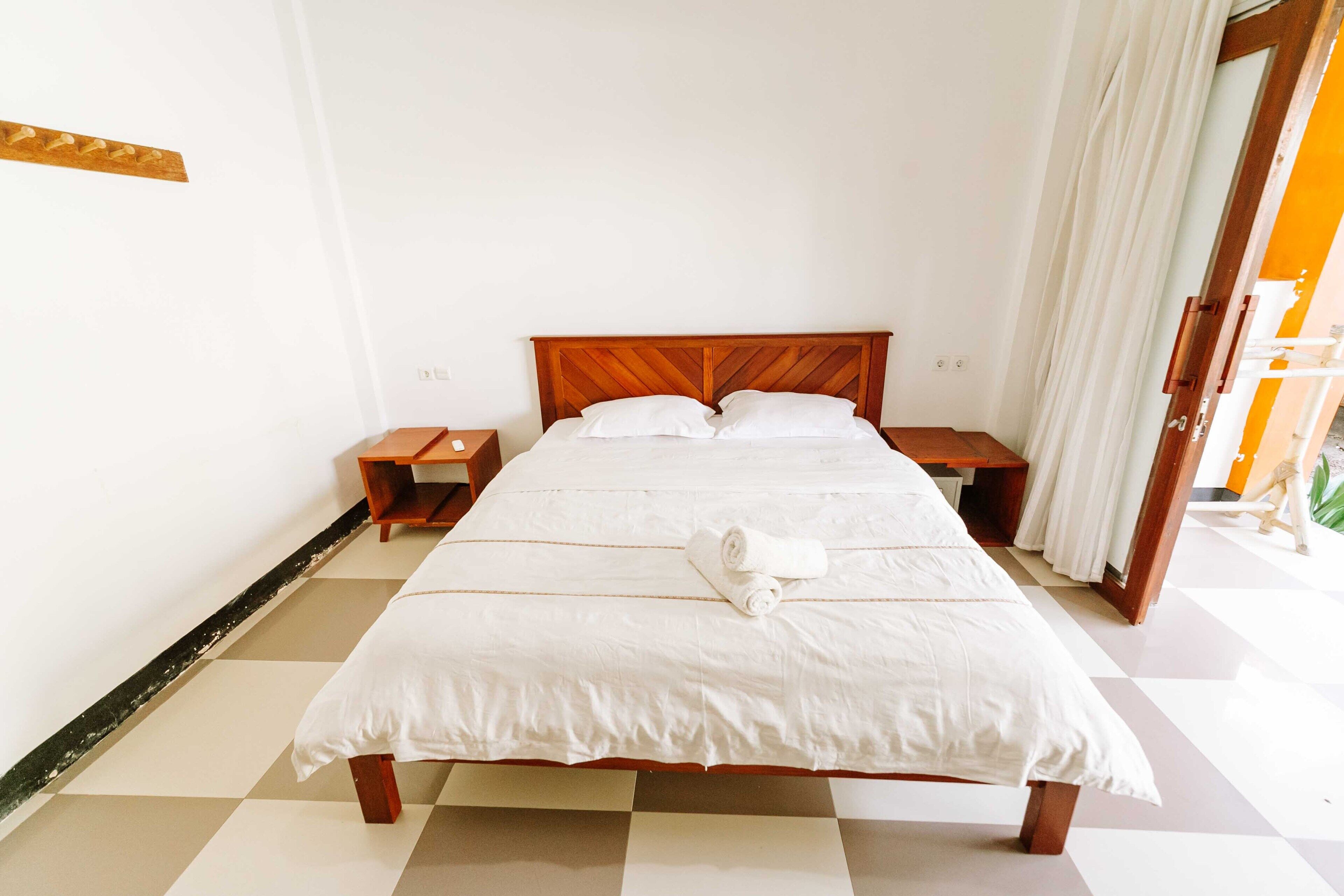 A neatly made bed with white linens and a wooden headboard, two bedside tables, and a curtain on the door.