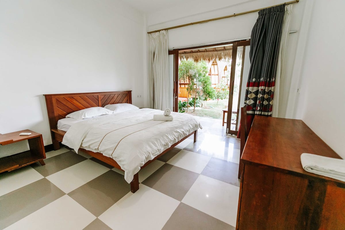 A bedroom with a wooden bed, a nightstand, a desk, and a view of a courtyard.