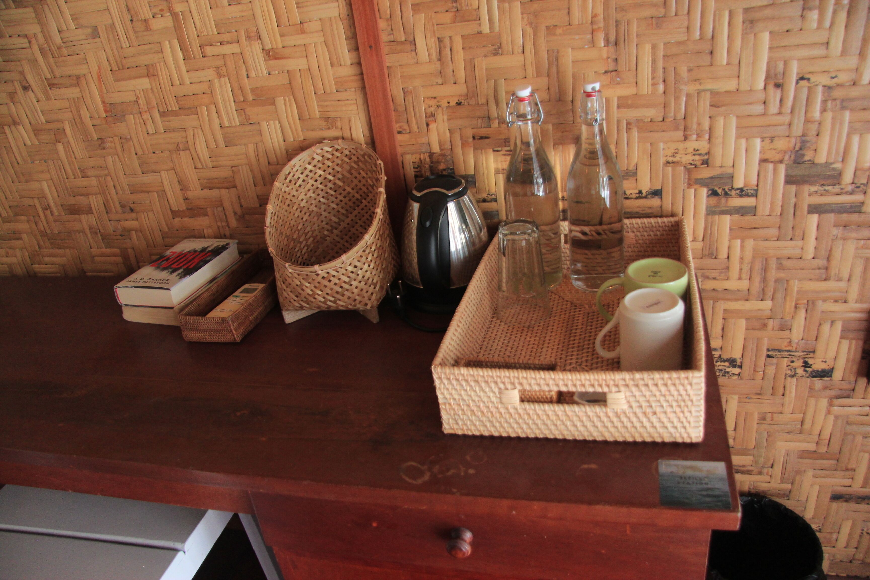 Una mesa de madera con cestas tejidas, una tetera, botellas y una bandeja con una taza y una vela.