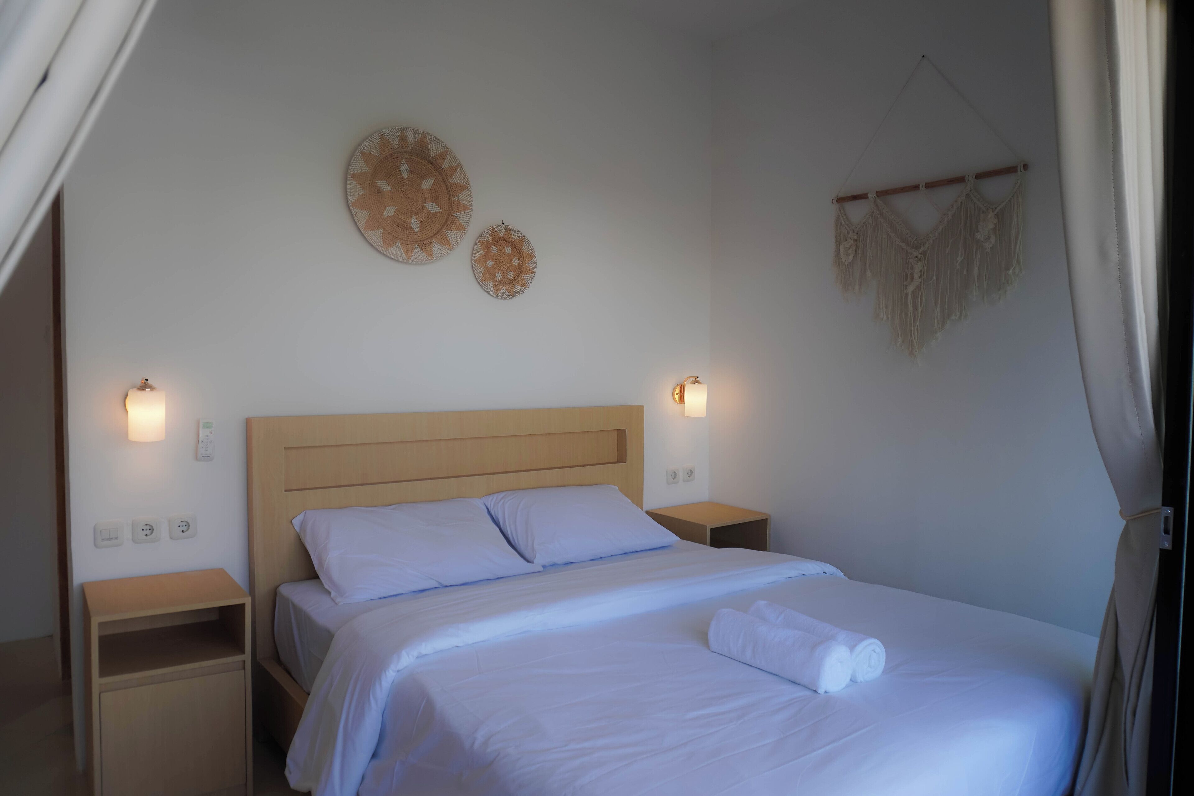 A neatly made bed with white linens and a rolled towel, a wooden headboard, bedside tables with lamps, and wall decor.