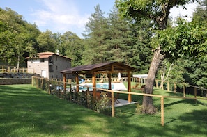 Una cabaña rústica con fachada de piedra y terraza de madera, rodeada de vegetación exuberante y un área de piscina vallada.